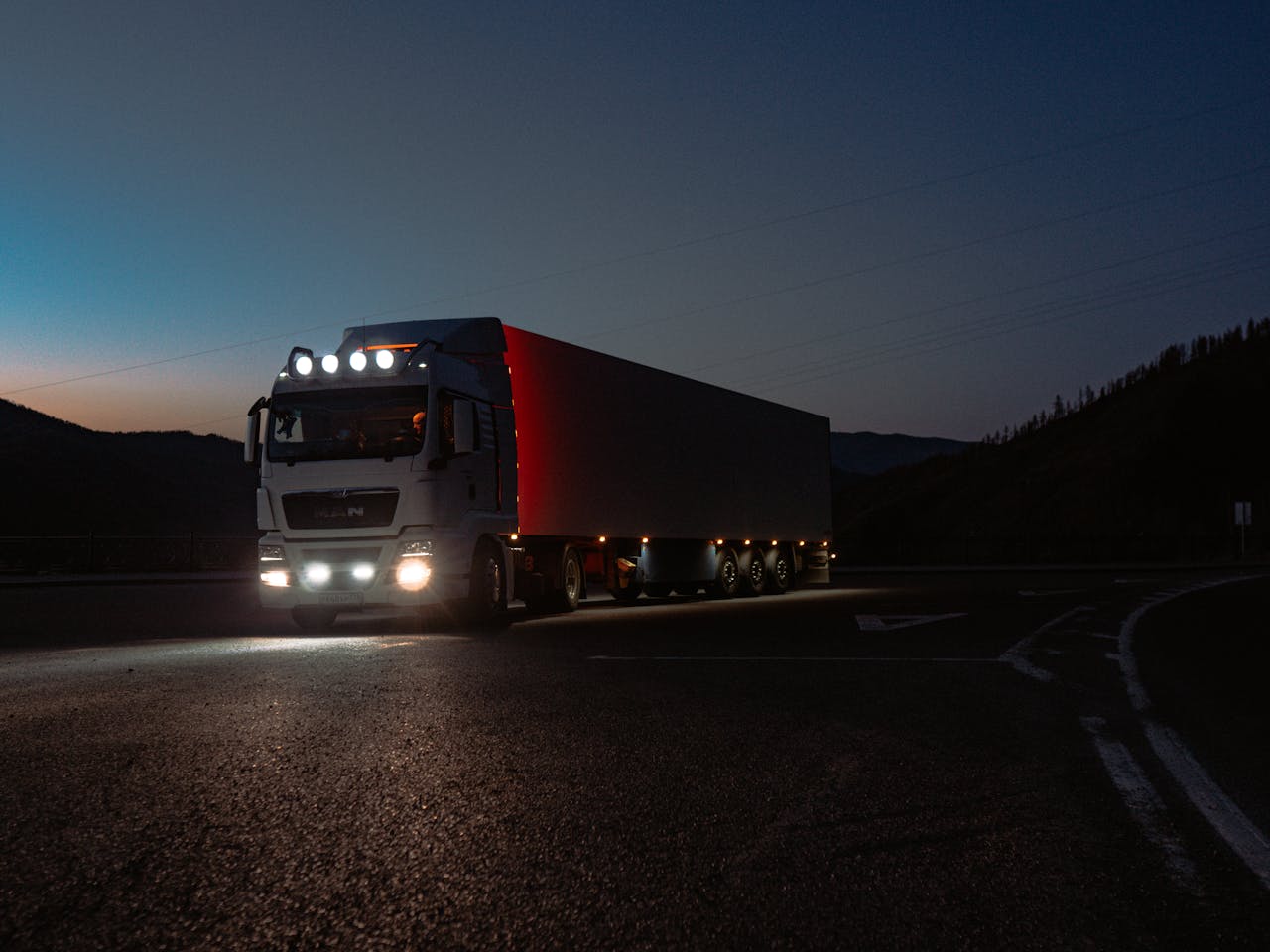 about-img A heavy-duty truck navigates a quiet road at dusk, highlighting its illuminated path through remote mountains.